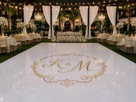 Elegant white vinyl wrapped dance floor featuring a custom gold "K & M" monogram. It is set in a beautiful outdoor wedding reception area decorated with string lights and white drapery